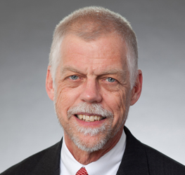 Smiling older man with short gray hair and beard, wearing a dark suit, white shirt, and red patterned tie—posed in front of a plain gray background—reflecting professionalism typical of leading Chicago lawyers in a corporate law office.