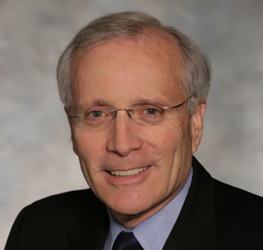 An older man with gray hair, wearing glasses, a dark suit, and a blue shirt, poses in front of a neutral, blurred background—reflecting the professionalism of a corporate law office.