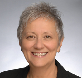 Smiling older woman with short gray hair wearing a black blazer, photographed against a plain light gray background, exuding the professionalism of a corporate law office.