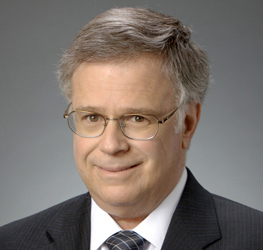 A middle-aged man with gray hair and glasses, dressed in a dark suit, striped tie, and light shirt, poses against a plain gray background—reflecting the professionalism found in leading corporate law office environments.
