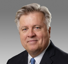 A middle-aged man with gray hair wearing a dark suit, white shirt, and blue tie, poses against a plain gray background—professional attire common among Chicago lawyers or those working in law offices.
