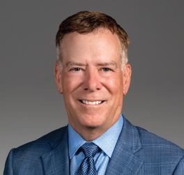Man in a blue suit and tie smiling at the camera against a plain gray background, representing professionalism often found at top law offices and among experienced lawyers in Chicago.
