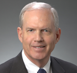 A middle-aged man with short gray hair, wearing a dark suit, white shirt, and blue tie, poses against a plain gray background—reflecting the professionalism of a corporate law office specializing in intellectual property law.