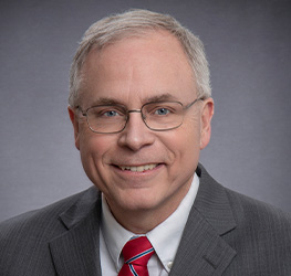 A middle-aged man with short gray hair, glasses, and a gray suit with a red striped tie, smiles at the camera against a plain gray background, reflecting the professionalism of a corporate law office.