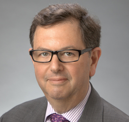 A middle-aged man with short brown hair, glasses, and a suit with a striped shirt and purple patterned tie poses confidently against a gray background, reflecting the professional image of corporate law office environments.
