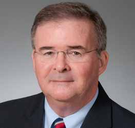 Middle-aged man with short brown hair, glasses, and a suit jacket over a light blue shirt and striped tie, posing against a plain gray background—perfect for a corporate law office or intellectual property law profile.