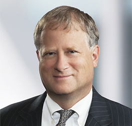 A middle-aged man with short, light brown hair is wearing a dark pinstripe suit, white shirt, and patterned tie, posing against a blurred professional background at one of the top law offices specializing in intellectual property law.