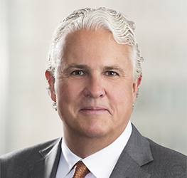 A middle-aged man with white hair wearing a gray suit, white shirt, and orange tie poses in front of a blurred light background, representing law offices specializing in intellectual property law.