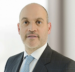 A bald man with a trimmed beard wearing a dark suit, white shirt, and light blue patterned tie, posing in front of a blurred light background—reflecting the professional image often seen at leading Chicago lawyers' law offices.