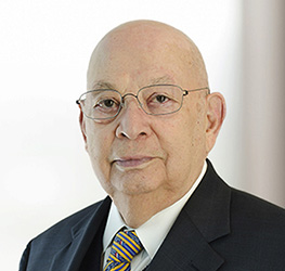 An older man with glasses, dressed in a dark suit, white shirt, and yellow-and-blue striped tie, faces the camera with a neutral expression—reflecting the professionalism found in top corporate law offices.