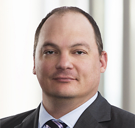 A man with short, thinning hair wears a dark suit, light blue shirt, and striped tie, posing in front of a blurred light-colored background—typical professional attire for lawyers in Chicago.