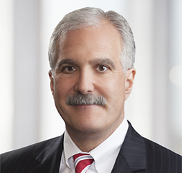 Middle-aged man with gray hair and mustache wearing a dark pinstripe suit, white shirt, and red striped tie, posed in front of a blurred corporate law office background.