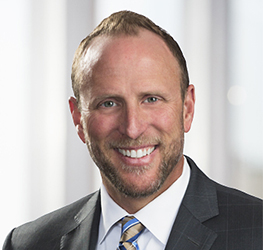 A man in a suit and tie smiles at the camera in a professional setting, possibly one of the law offices specializing in intellectual property law, with a blurred light-colored background.