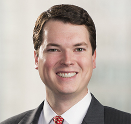A man in a dark suit, white shirt, and red patterned tie smiles at the camera against a blurred light background, reflecting the professionalism of chicago lawyers in a leading corporate law office.