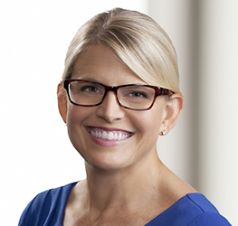 Smiling woman with blonde hair, wearing glasses and a blue top, stands in front of a light-colored background—ideal for a corporate law office specializing in intellectual property law.