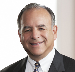 A middle-aged man with short gray hair, wearing a dark suit, white shirt, and patterned tie, smiles in a professional law offices setting.
