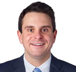 Man with short dark hair wearing a navy suit, white shirt, and light blue tie, smiling at the camera against a plain white background, representing a professional from a corporate law office.