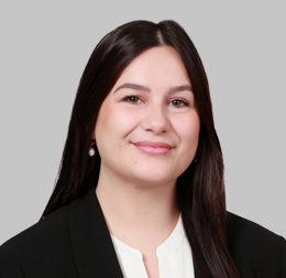 Woman with long dark hair wearing a black blazer and white blouse, smiling in front of a plain gray background, suited for a professional at a corporate law office.