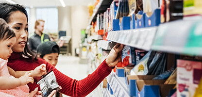 A woman holds a toddler while browsing products on a supermarket shelf with a young child beside her, perhaps picking up snacks after visiting lawyers in Chicago.