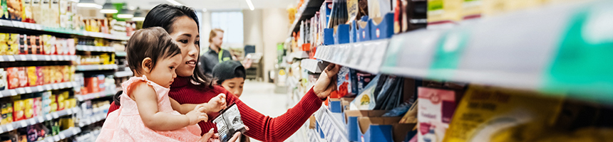 A woman holds a toddler while browsing products on a supermarket shelf with a young child beside her, perhaps picking up snacks after visiting lawyers in Chicago.