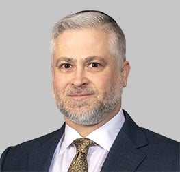 A middle-aged man with short gray hair and a beard, dressed in a dark suit, white shirt, and patterned tie, poses against a plain light gray background—reflecting the professionalism of lawyers in Chicago at a leading corporate law office.