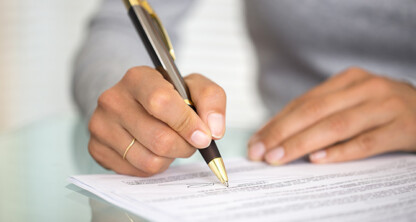 At a corporate law office, a person holding a pen signs a document on a glass table.