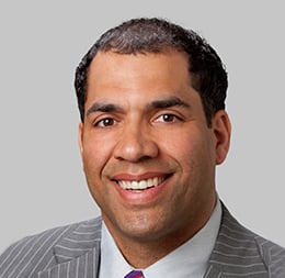 A man in a gray pinstripe suit, white shirt, and purple tie smiles at the camera against a plain light gray background, reflecting the professionalism found in top corporate law offices.