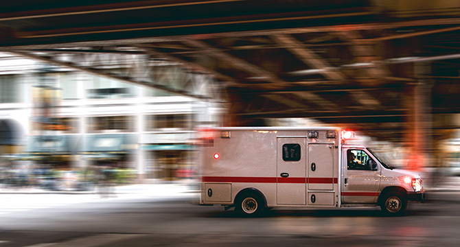 An ambulance with flashing lights drives quickly under an overpass in an urban area, passing by law offices offering litigation support.
