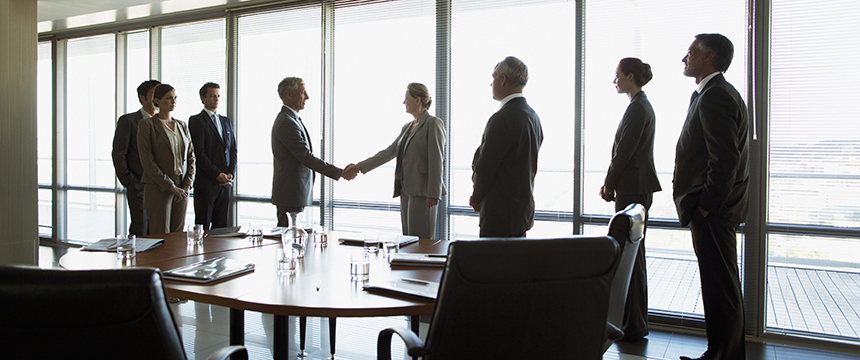 Seven business professionals stand in a corporate law office conference room, with two in the center shaking hands across a table, while others observe.