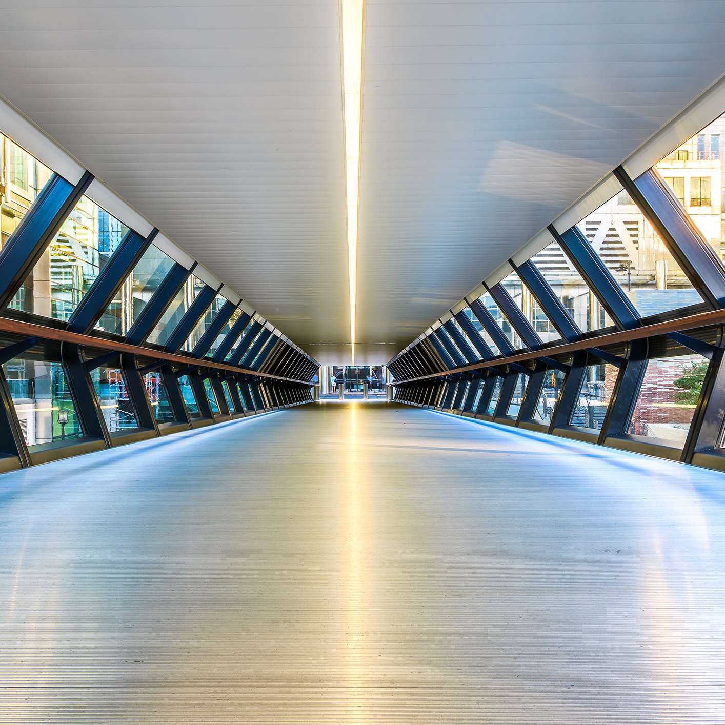 Modern pedestrian skybridge with glass walls and a metal floor, leading to an urban area where a corporate law office is visible among the buildings at the far end.
