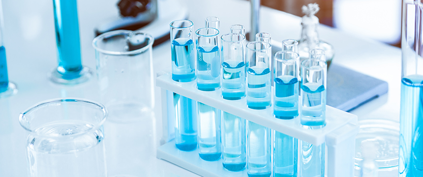 A set of test tubes with blue liquid arranged in a white rack on a laboratory table, surrounded by other glassware, evokes the precision often found in intellectual property law offices.