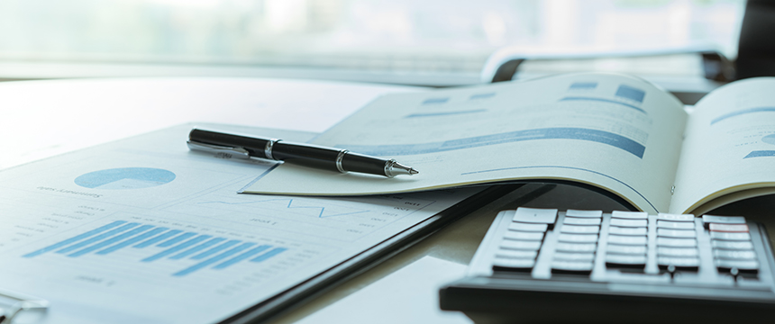 A pen rests on an open report with charts and graphs, beside a calculator and more financial documents on the desk of a corporate law office.