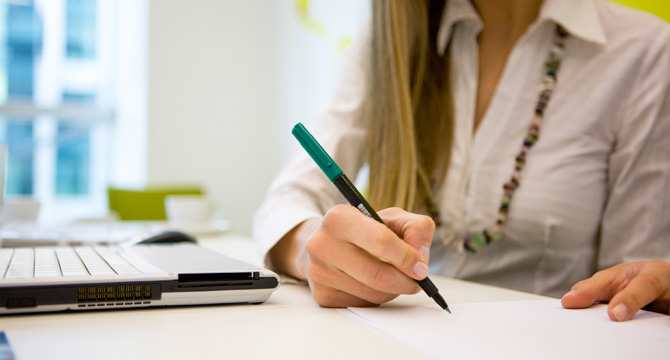 A person in a white shirt writes on paper at a desk beside an open laptop, perhaps researching intellectual property law or preparing documents for Chicago lawyers.