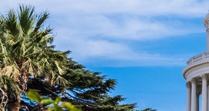 California State Capitol dome with U.S. and California flags at half-mast, palm trees, blue sky, and a corporate law office nearby specializing in intellectual property law.