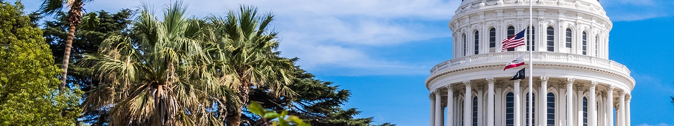 California State Capitol dome with U.S. and California flags at half-mast, palm trees, blue sky, and a corporate law office nearby specializing in intellectual property law.