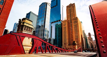 A red steel bridge spans a city street, with tall skyscrapers and historic law offices under a partly cloudy sky in the background.