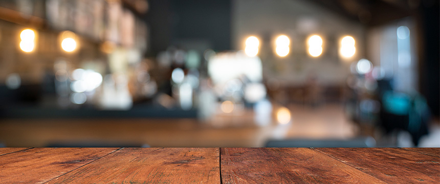 A wooden table in the foreground with a blurred view of a café or restaurant interior resembling modern law offices in the background, featuring lights and furniture.