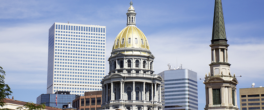 A view of the Colorado State Capitol with its gold dome, surrounded by modern office buildings, a corporate law office, and a church steeple, under a partly cloudy sky.