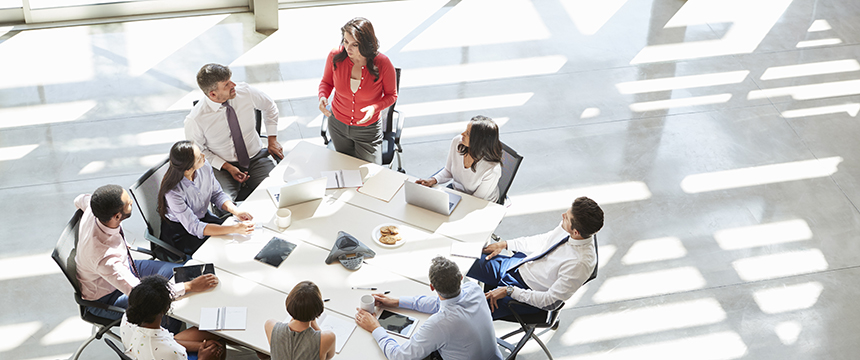 Eight people sit around a table in a modern corporate law office, engaged in a meeting; one woman stands and speaks while others listen and take notes.