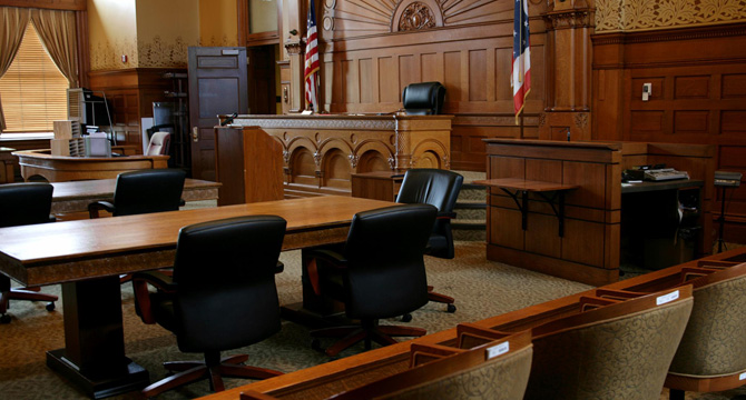 A courtroom with empty tables, chairs, a judge's bench, flags, and wooden decor. The atmosphere evokes the seriousness found in litigation support for complex cases or corporate law office proceedings. No people are present.
