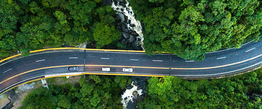 Aerial view of a curved road with three cars driving through dense green forest over a small river—an inspiring scene for lawyers in Chicago seeking tranquility outside the city.