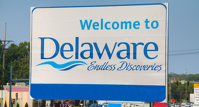 A road sign reads "Welcome to Delaware Endless Discoveries" with a blue wave graphic, set against a backdrop of trees, buildings, and a traffic light—a welcome sight for those seeking intellectual property law expertise.