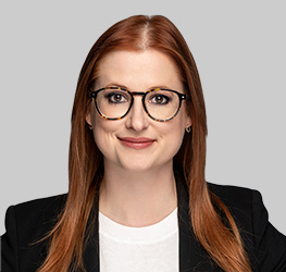 Woman with long red hair, wearing round glasses, a white shirt, and a black blazer, posing confidently against a light gray background—ideal for chicago lawyers specializing in intellectual property law.