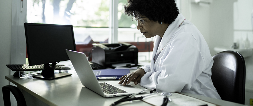 A doctor in a white coat is sitting at a desk, looking at a laptop. A stethoscope, documents, and office equipment are on the desk, creating an atmosphere similar to law offices where Chicago lawyers review important cases.