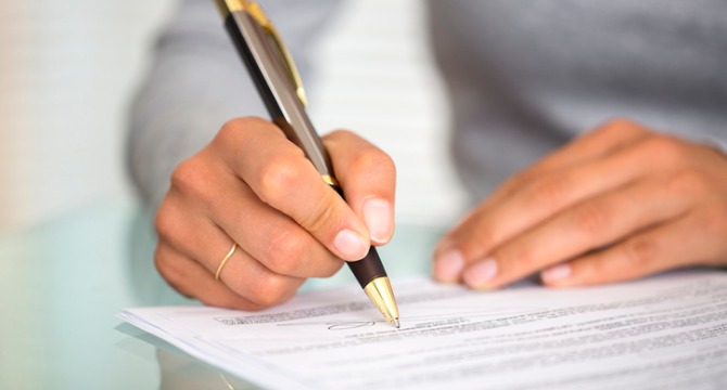 A person holding a pen signs a document on a glass table, with text visible on the paper—a scene commonly found in law offices or with lawyers in Chicago.