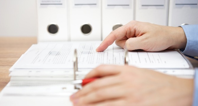 A person points to a page in an open binder filled with documents, with several closed binders arranged on a shelf in the background—typical of busy law offices specializing in intellectual property law.