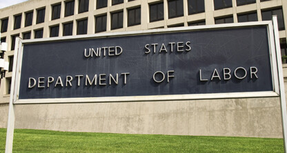 A sign reading "United States Department of Labor" stands in front of a large government building with rows of windows and a grassy lawn, reminiscent of the impressive presence found at a major corporate law office.