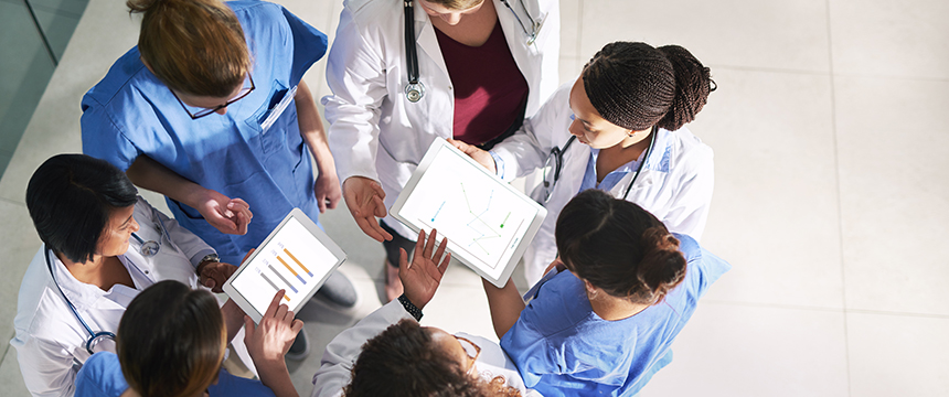 A group of healthcare professionals, wearing scrubs and lab coats, stand in a circle at a corporate law office and review data on digital tablets.