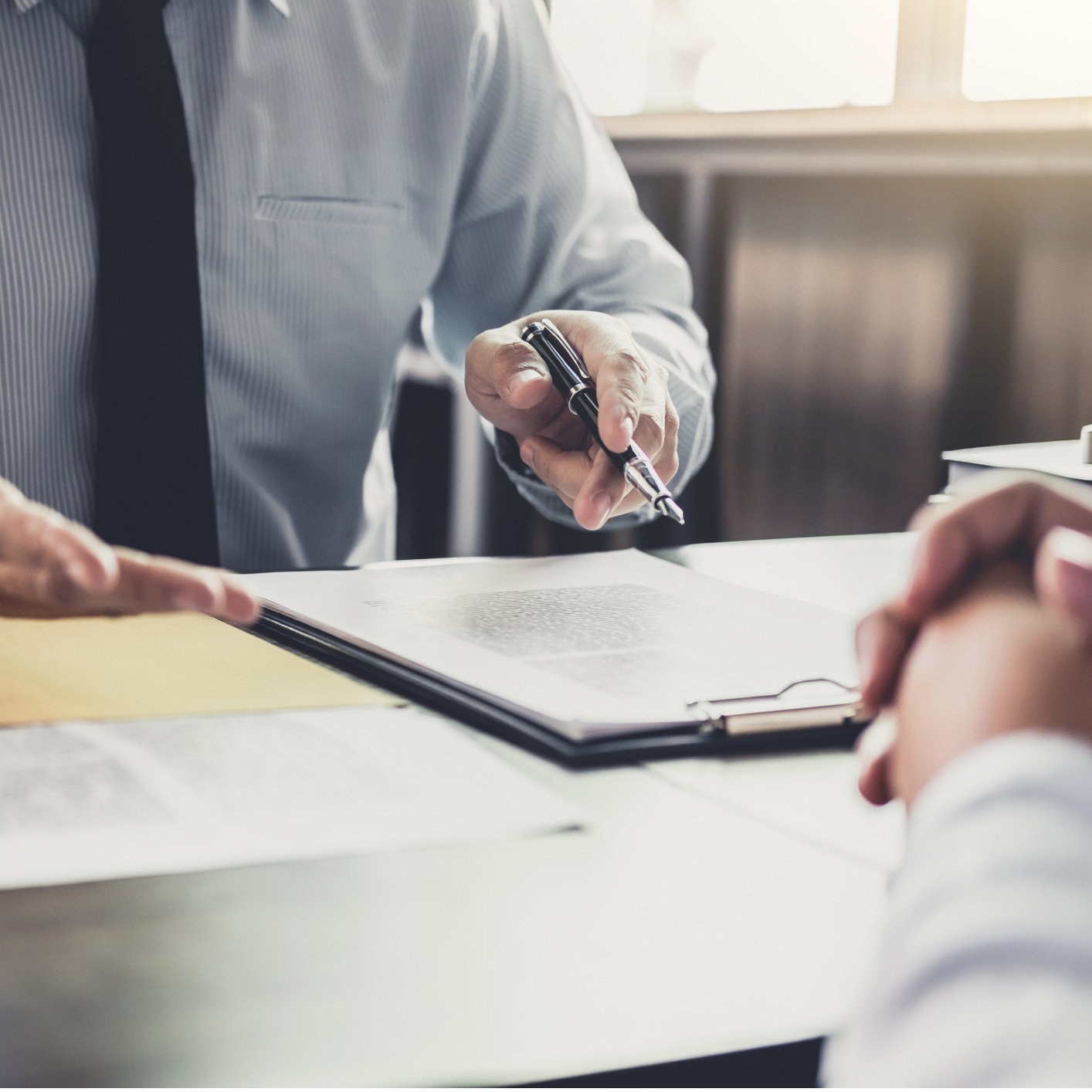 Two people sit at a desk reviewing and discussing documents; one person points with a pen at a clipboard holding papers, illustrating the collaborative process often found in law offices.