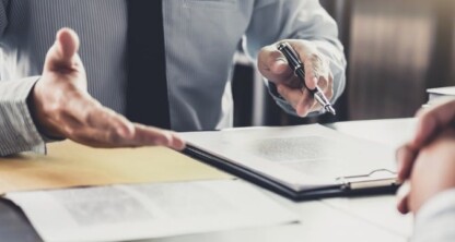 Two people sit across a desk in a corporate law office, one holding a pen and explaining something while pointing to a document on a clipboard.